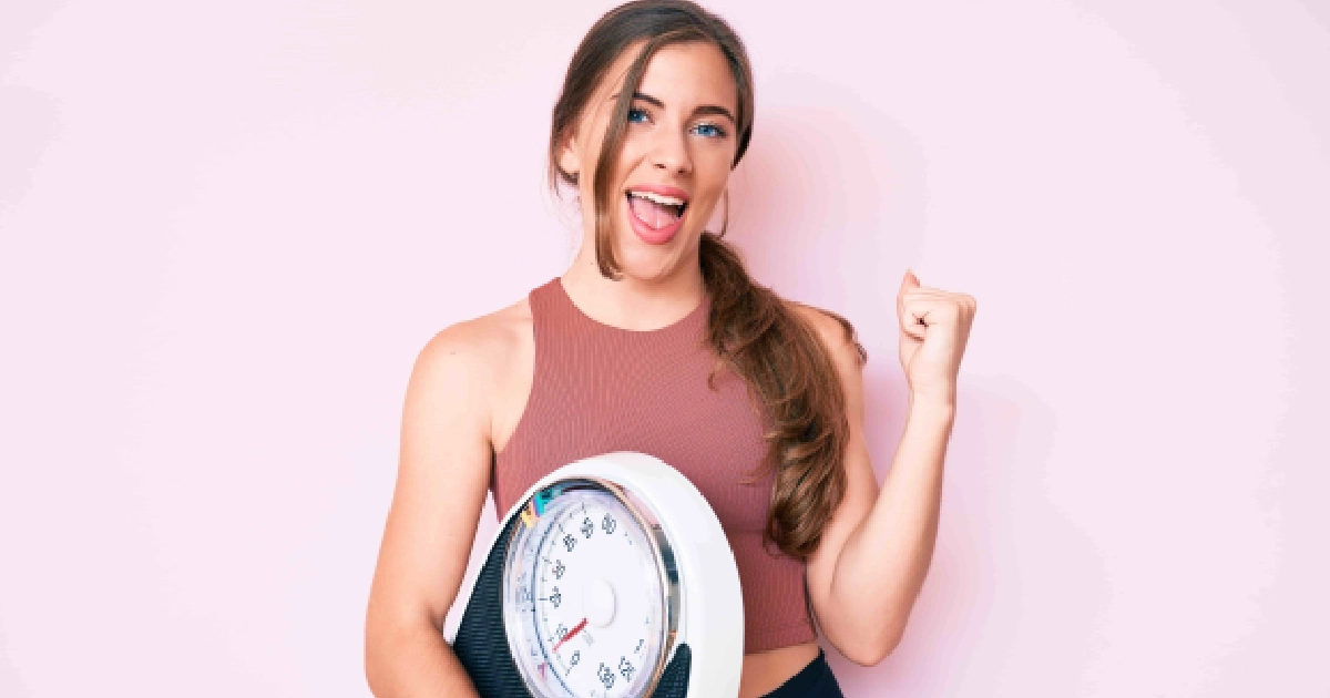 Smiling woman in a pink crop top holding a bathroom scale and celebrating against a light pink background, representing Sustainable Weight Management in St. Peters, MO.