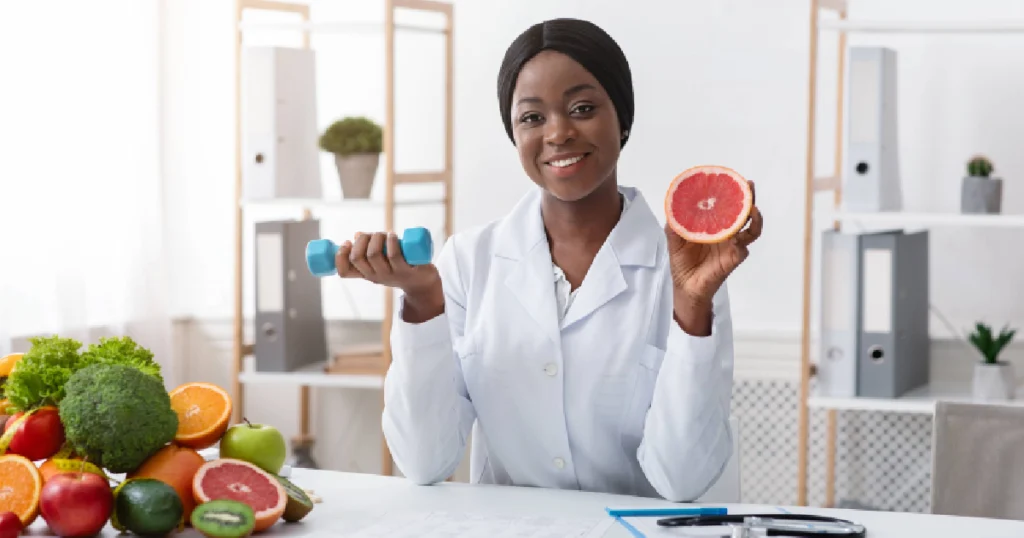 Smiling female doctor holding a fruit and dumbbell at her desk surrounded by fresh fruits, representing holistic Wellness Treatments in St. Peters, MO.